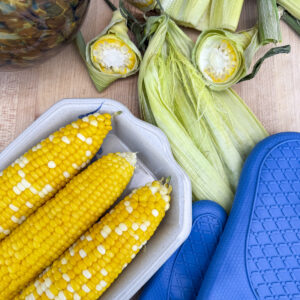 three cooked cobs of corn on plate completely free of silk, next to blue oven mitts and corn husk with silk