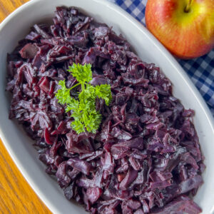 braised red cabbage - dark purple color on white serving dish with sprig of parsley and a fresh apple beside it