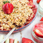 Bright red strawberry sits on top of strawberry rhubarb crisp. Rhubarb and strawberries surround.